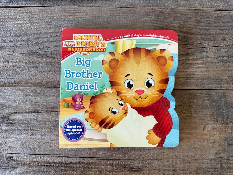 Puede incluir: Un libro infantil titulado "Big Brother Daniel" de Daniel Tiger's Neighborhood. La portada presenta una ilustraci&oacute;n de Daniel Tiger sosteniendo un beb&eacute; tigre. El libro tiene un borde rojo y azul y el texto "Based on the special episode!"