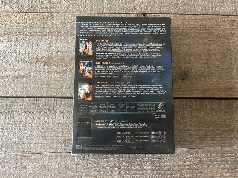 K&ouml;nnte beinhalten: Ein DVD-Box-Set mit der Die Hard-Film-Trilogie. Die Box ist schwarz mit silbernem Rand und zeigt Bilder von Bruce Willis als John McClane aus jedem Film. Die R&uuml;ckseite der Box enth&auml;lt eine Beschreibung jedes Films und eine Liste der besonderen Merkmale.