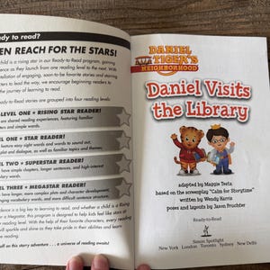 May include: An open book with the title "Daniel Visits the Library" featuring Daniel Tiger characters. The book is part of the Ready-to-Read series, with text about reading levels and a program to encourage children to read.
