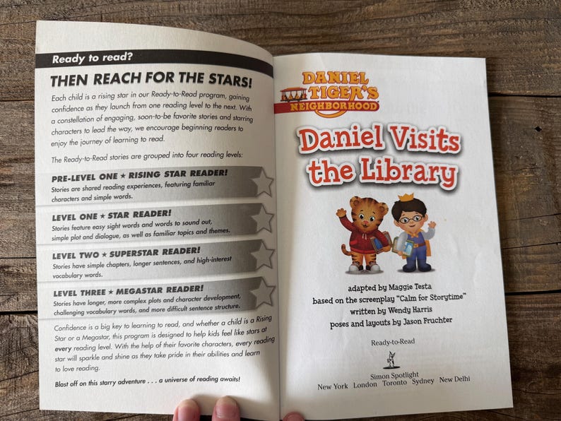 May include: An open book with the title "Daniel Visits the Library" featuring Daniel Tiger characters. The book is part of the Ready-to-Read series, with text about reading levels and a program to encourage children to read.
