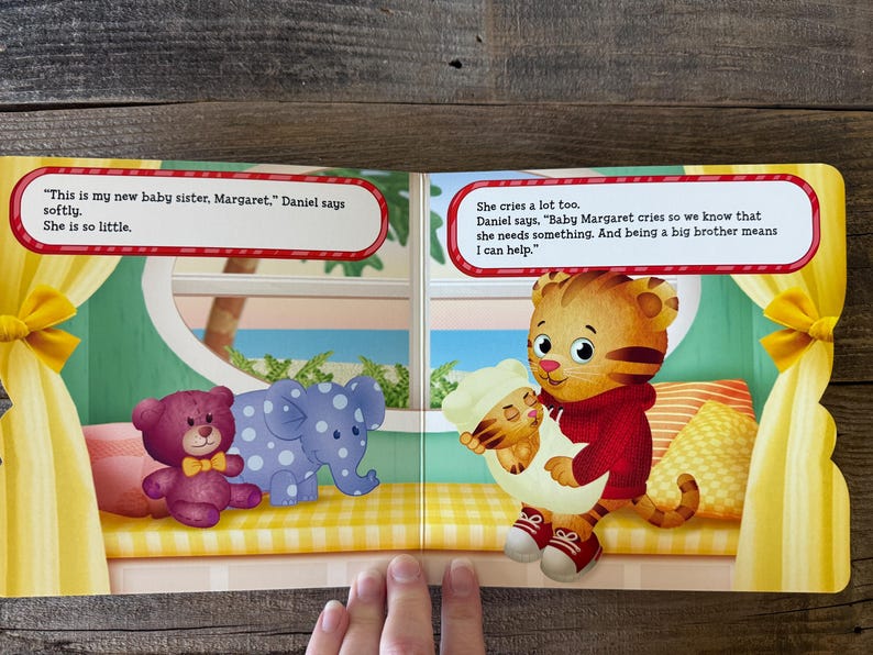 Puede incluir: Doble p&aacute;gina de un libro infantil con Daniel Tigre sosteniendo un beb&eacute; tigre. A la izquierda, un oso de peluche morado y un elefante azul con lunares blancos. Las burbujas de texto contienen di&aacute;logo.