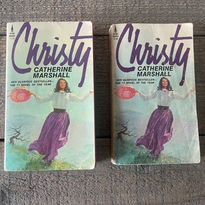 Pode incluir: Duas cópias de bolso do romance "Christy" de Catherine Marshall. A capa apresenta uma mulher com um vestido roxo e uma blusa branca, segurando um chapéu. O texto "HER GLORIOUS BESTSELLER - THE #1 NOVEL OF THE YEAR" está impresso na capa.