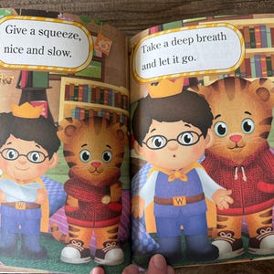 May include: A children's book illustration featuring two cartoon characters, one a boy wearing glasses and a crown, and the other a tiger. Text bubbles read "Give a squeeze, nice and slow" and "Take a deep breath and let it go."
