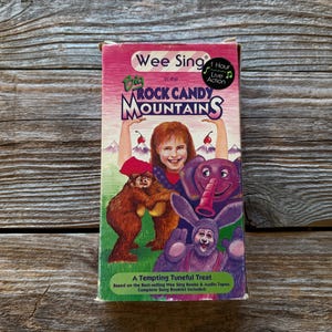 May include: A vintage VHS tape for "Wee Sing in the Big Rock Candy Mountains." The cover features a smiling girl with cartoon characters, including a bear, elephant, and rabbit. The tape is on a wooden surface.