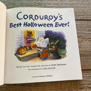 May include: A book cover featuring a brown bear dressed in a red robe holding a plate of cookies. The bear is standing in front of a house with a pumpkin on the porch. The book title is "Corduroy's Best Halloween Ever!" and the text below the title reads "Based on the character created by Don Freeman Illustrated by Lisa McCue Grosset & Dunlap, Publishers."
