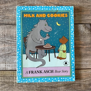 Puede incluir: Un libro infantil titulado "Milk and Cookies" con un borde azul. La portada presenta un dragón gris y un oso pardo, ambos cerca de una mesa con galletas. El texto "A FRANK ASCH Bear Story" está en la parte inferior.