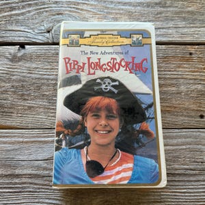 May include: A VHS tape of "The New Adventures of Pippi Longstocking" from the Columbia TriStar Family Collection. The cover features a smiling girl with red hair wearing a pirate hat. The background shows a ship and blue sky.