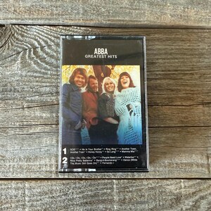 以下が含まれることがあります： ABBAの「Greatest Hits」のヴィンテージカセットテープ。カバーにはバンドメンバーのカラー写真が写っています。「Dancing Queen」や「Waterloo」などの曲が収録されています。カセットは木製の背景に置かれています。