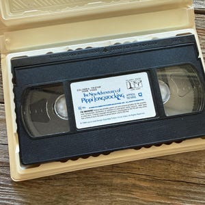 May include: A black VHS tape of "The New Adventures of Pippi Longstocking" in a beige plastic case. The tape has a white label with the movie title and a logo. The case is open, revealing the tape inside.