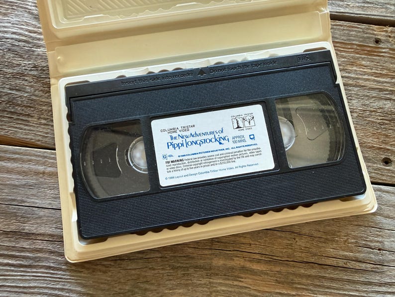 May include: A black VHS tape of "The New Adventures of Pippi Longstocking" in a beige plastic case. The tape has a white label with the movie title and a logo. The case is open, revealing the tape inside.