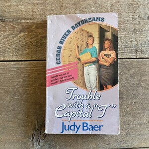 Könnte beinhalten: Ein rosa-weißes Taschenbuch mit dem Titel "Trouble with a "T" Capital "T" by Judy Baer". Das Cover zeigt ein Foto von zwei Mädchen in einem Schulflur mit Schließfächern im Hintergrund.
