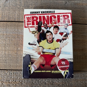 Peut inclure: Boîtier DVD du film "The Ringer" avec Johnny Knoxville. La couverture montre un groupe d'hommes dans diverses poses, avec le titre en grandes lettres rouges. Le boîtier comprend le texte "Laugh-Out-Loud Hilarious" et "15 Minutes of Outrageous Deleted Scenes."