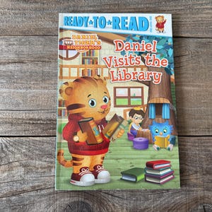 May include: A children's book titled "Daniel Visits the Library" featuring Daniel Tiger holding books. The cover has a colorful illustration of Daniel Tiger, a library scene, and the words "Ready to Read".