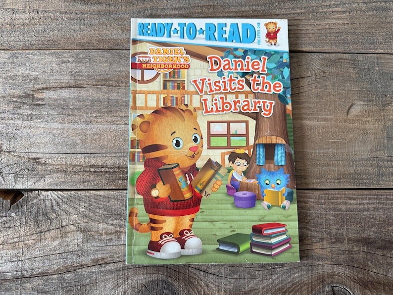 May include: A children's book titled "Daniel Visits the Library" featuring Daniel Tiger holding books. The cover has a colorful illustration of Daniel Tiger, a library scene, and the words "Ready to Read".