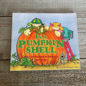 Può includere: Una copertina di libro con un'illustrazione di due bambini e un cane all'interno di una grande zucca arancione. Il titolo del libro è "In a Pumpkin Shell" e il sottotitolo è "Over 20 Pumpkin Projects for Kids". Il libro è di Jennifer Storey Gillis.
