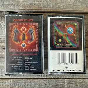 May include: Two vintage Journey "Greatest Hits" cassette tapes. The first features a red and gold design with a winged figure. The second has a colourful, rainbow-themed design with a planet. Both are on a wooden surface.
