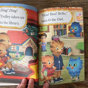 May include: A children's book spread featuring illustrations of Daniel Tiger and friends. Text bubbles read "Ding! Ding! Trolley takes us to the library" and "Hoo! Hoo! Hello, says O the Owl."