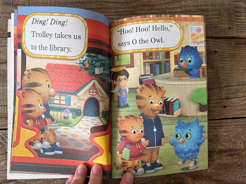 May include: A children's book spread featuring illustrations of Daniel Tiger and friends. Text bubbles read "Ding! Ding! Trolley takes us to the library" and "Hoo! Hoo! Hello, says O the Owl."