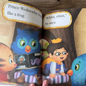 May include: A children's book spread featuring illustrations of cartoon characters. Text bubbles read "Prince Wednesday hops like a frog" and "Ribbit, ribbit, he says." The characters include a prince, an owl, a cat, and a blue bird.
