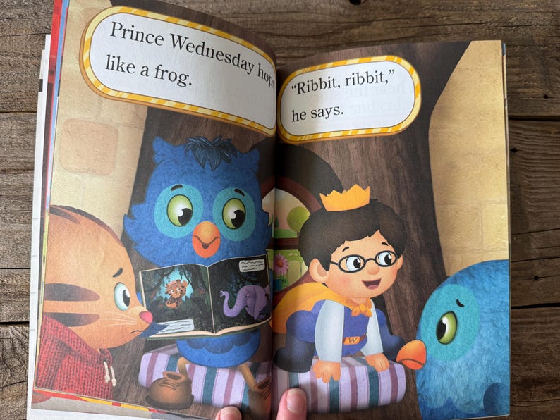 May include: A children's book spread featuring illustrations of cartoon characters. Text bubbles read "Prince Wednesday hops like a frog" and "Ribbit, ribbit, he says." The characters include a prince, an owl, a cat, and a blue bird.