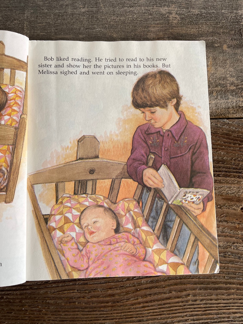 Pu&ograve; includere: Un bambino legge un libro alla sua sorellina che dorme in una culla. La culla ha una trapunta colorata. Il bambino indossa una camicia viola e jeans. Il testo nell'immagine dice: "A Bob piaceva leggere. Ha cercato di leggere alla sua nuova sorella e mostrarle le immagini nei suoi libri. Ma Melissa ha sospirato e ha continuato a dormire."