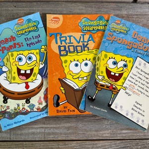 Może przedstawiać: Trzy kolorowe książki SpongeBob Kanciastoporty są wyświetlane na drewnianej powierzchni. Tytuły to "The Last Episode", "Trivia Book" i "Dear SpongeBob". Każda książka zawiera ilustracje i tekst SpongeBoba, z żywymi niebieskimi, pomarańczowymi i żółtymi okładkami.
