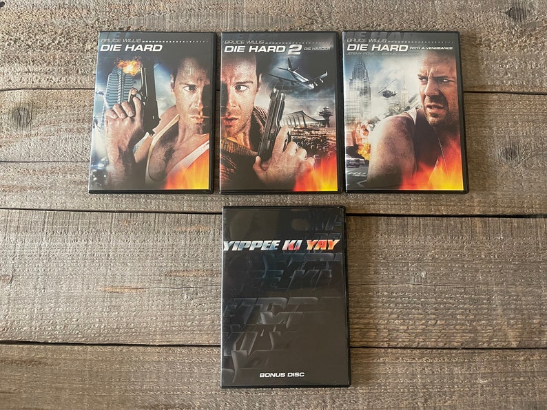 K&ouml;nnte beinhalten: Vier DVD-H&uuml;llen mit schwarzen Covern und rotem und wei&szlig;em Text. Die Titel sind "Die Hard", "Die Hard 2", "Die Hard With a Vengeance" und "Yippee Ki Yay" mit den Worten "Bonus Disc" am unteren Rand des letzten Geh&auml;uses.