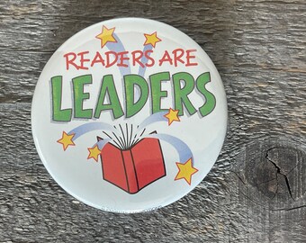 Könnte beinhalten: Ein weißer Knopf mit einem roten Buch, blau-weißen Feuerwerkskörpern und dem Text "Readers Are Leaders" in grünen Buchstaben mit gelben Sternen.