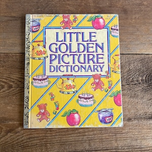 Puede incluir: Un libro Little Golden Book amarillo, azul y blanco con el título "Little Golden Picture Dictionary". La portada presenta ilustraciones de una tetera, un osito de peluche, un pastel y una manzana.