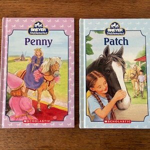 Könnte beinhalten: Zwei Taschenbücher für Kinder mit rosa und blauen Umschlägen. Das Buch links zeigt ein Mädchen, das ein braunes Pferd reitet, mit dem Titel "Penny", und das Buch rechts zeigt ein Mädchen, das ein schwarz-weißes Pferd streichelt, mit dem Titel "Patch". Beide Bücher gehören zur Breyer Stablemates-Reihe.