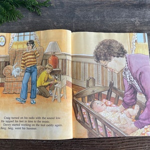 Pu&ograve; includere: Un libro aperto con un'illustrazione di una famiglia in una casa. Una donna tiene un bambino in una culla. Un ragazzo sta lavorando su un carrello porta attrezzi. Un altro ragazzo sta ascoltando musica alla radio. Il testo sulla pagina dice: "Craig ha acceso la sua radio con il volume basso. Ha battuto i piedi a tempo con la musica. Davey ha iniziato a lavorare sul carrello porta attrezzi di nuovo. Bang, bang, &egrave; andato il suo martello."