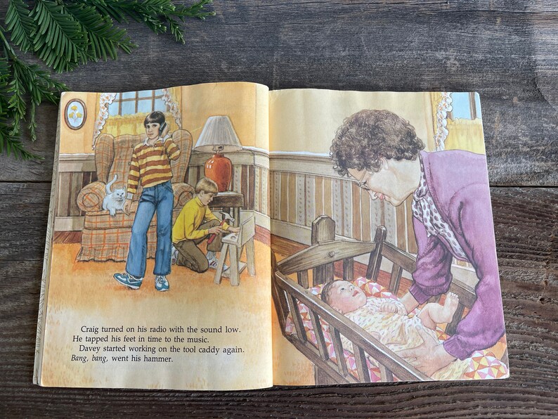 Pu&ograve; includere: Un libro aperto con un'illustrazione di una famiglia in una casa. Una donna tiene un bambino in una culla. Un ragazzo sta lavorando su un carrello porta attrezzi. Un altro ragazzo sta ascoltando musica alla radio. Il testo sulla pagina dice: "Craig ha acceso la sua radio con il volume basso. Ha battuto i piedi a tempo con la musica. Davey ha iniziato a lavorare sul carrello porta attrezzi di nuovo. Bang, bang, &egrave; andato il suo martello."