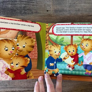 Puede incluir: Ilustraci&oacute;n de libro infantil con personajes de Daniel el Tigre. La p&aacute;gina izquierda muestra a una familia abraz&aacute;ndose, con el texto: &laquo;A veces un hermano mayor necesita un gran abrazo&raquo;. La p&aacute;gina derecha muestra a Daniel el Tigre con su familia.