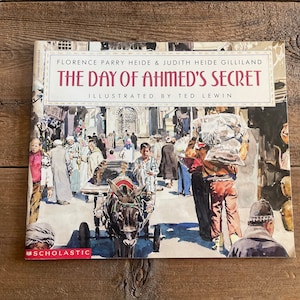 May include: A book cover with a watercolor illustration of a bustling marketplace in a Middle Eastern city. The title of the book is "The Day of Ahmed's Secret" by Florence Parry Heide and Judith Heide Gilliland, illustrated by Ted Lewin. The book is published by Scholastic.