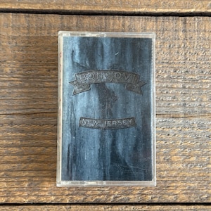 May include: A vintage cassette tape with a dark blue cover. The tape case features the band name "BON JOVI" in a banner and the text "NEW JERSEY" below. The cassette is set against a wooden background.