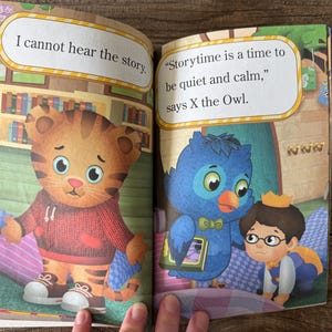 May include: A children's book spread featuring illustrations of cartoon characters. One page shows a tiger-like character with the text "I cannot hear the story." The other page shows an owl and a boy with the text "Storytime is a time to be quiet and calm."