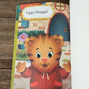 May include: A children's book page featuring a cartoon tiger wearing a red sweater. The tiger is smiling and has its arms outstretched. Above the tiger is a speech bubble with the words "Ugga Mugga!" The background includes a house and a flower.