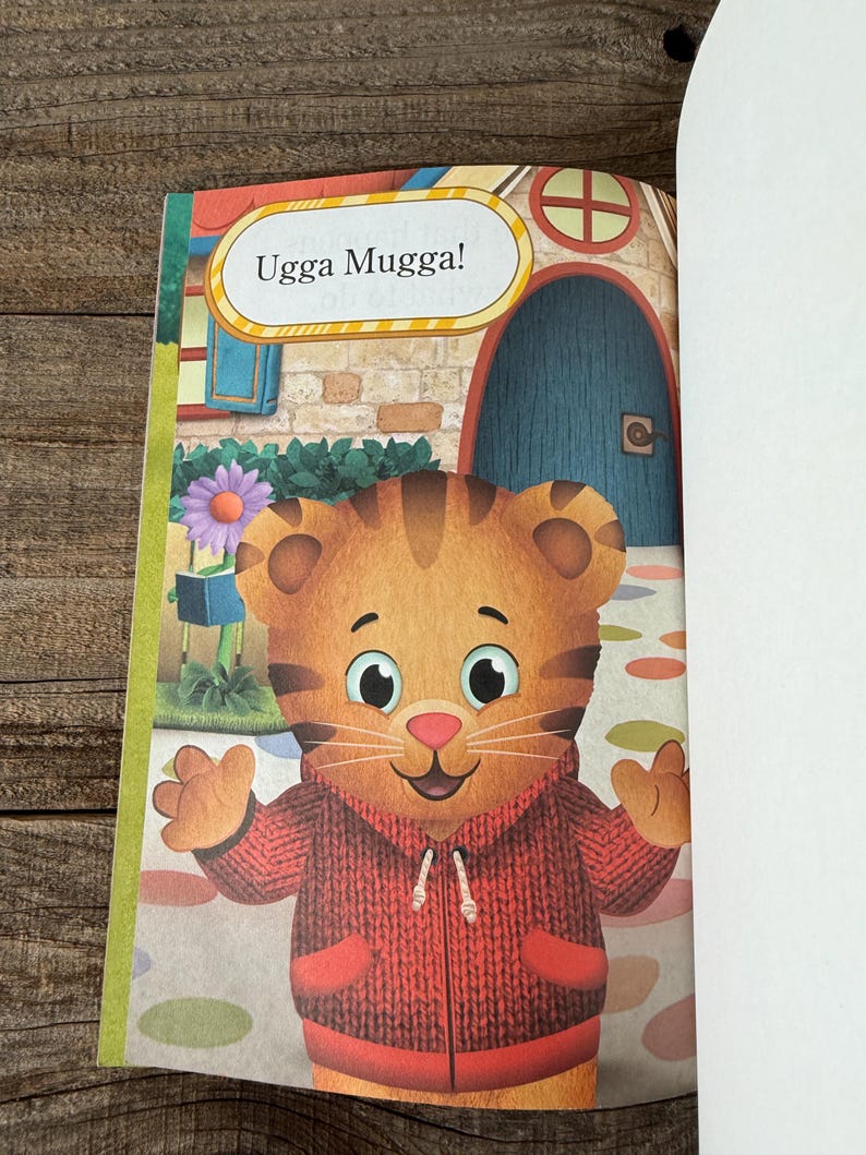 May include: A children's book page featuring a cartoon tiger wearing a red sweater. The tiger is smiling and has its arms outstretched. Above the tiger is a speech bubble with the words "Ugga Mugga!" The background includes a house and a flower.