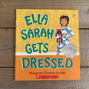 Puede incluir: Una cubierta de libro amarilla con un título verde que dice "Ella Sarah Gets Dressed". La cubierta presenta a una niña en pijama sosteniendo un animal de peluche. El libro está publicado por Scholastic.