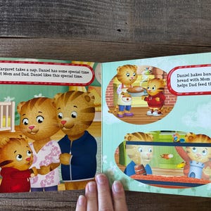 Puede incluir: Libro abierto con ilustraciones del programa infantil Daniel Tiger's Neighborhood. Las p&aacute;ginas muestran a Daniel Tiger con sus padres, y el texto dice: "Cuando Margaret toma una siesta, Daniel tiene un tiempo especial con mam&aacute; y pap&aacute;."