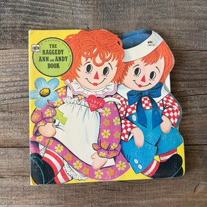以下が含まれることがあります： 「The Raggedy Ann and Andy Book」というタイトルのヴィンテージ子供向けの本。表紙には、ラガディ・アンとアンディの人形のイラストが描かれています。ラガディ・アンは花柄のドレスを着ており、アンディはオーバーオールを着ています。本は木製の表面にあります。