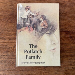 May include: A hardback book titled "The Potlatch Family" by Evelyn Sibley Lampman. The cover shows an illustration of a group of people and a woman's profile in muted tones. The book is on a wooden surface.