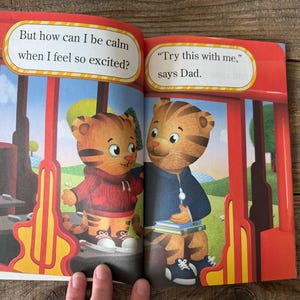 May include: Open book with illustrations of Daniel Tiger characters. One character wears a red hoodie, the other a blue jacket. Text bubbles read, "But how can I be calm when I feel so excited?" and "Try this with me," says Dad."