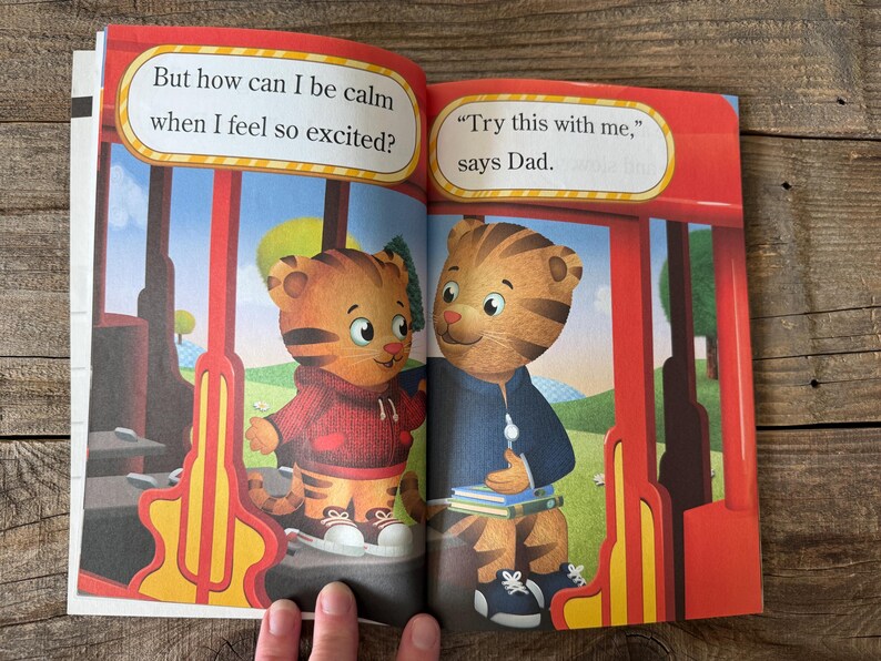 May include: Open book with illustrations of Daniel Tiger characters. One character wears a red hoodie, the other a blue jacket. Text bubbles read, "But how can I be calm when I feel so excited?" and "Try this with me," says Dad."