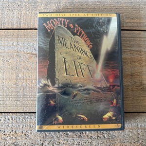 May include: A DVD case for "Monty Python's The Meaning of Life" with a tombstone graphic. The case is a two-disc special edition, with the word "Widescreen" at the bottom. The background is a wooden surface.
