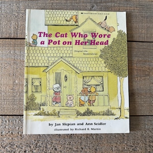 May include: A vintage children's book cover with a yellow house and a cat wearing a pot on its head. The title of the book is "The Cat Who Wore a Pot on Her Head" with the original title "Bendemelena" in parentheses. The book is by Jan Slepian and Ann Seidler, illustrated by Richard E. Martin.