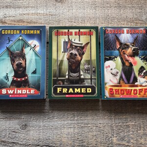 May include: Three paperback books by Gordon Korman featuring a dog on the cover. The titles are "Swindle", "Framed", and "Showoff".