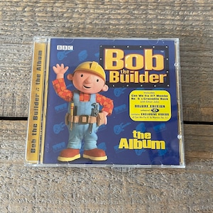 Könnte beinhalten: Ein CD-Gehäuse mit Bob der Baumeister, einer Zeichentrickfigur, die einen gelben Helm und ein blau-rotes Hemd trägt. Das Gehäuse ist blau und gelb und trägt den Text "Bob der Baumeister Das Album". Das Album enthält die Songs "Can We Fix It?", "Mambo No. 5" und "Crocodile Rock".