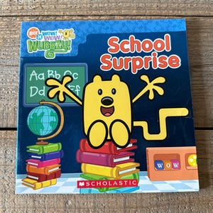 May include: A yellow cartoon character with big arms and legs is standing on a stack of books. The character is from the Nick Jr. show "Wow! Wow! Wubbzy!" The book cover has the title "School Surprise" and is published by Scholastic.