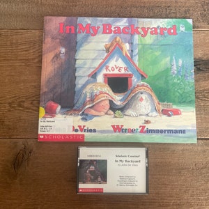 May include: A Scholastic Cassette tape with a colorful illustration of a child and a dog hiding under a blanket in a backyard. The cassette tape is titled "In My Backyard" by John De Vries.